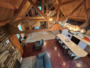 A spacious loft-style room with a high ceiling, exposed wooden beams, a cozy seating area with a sofa and chairs, a kitchen with modern appliances, and a view of the upper floor. - Private, cozy cabin on 13 acres with two ponds (Bloomfield)