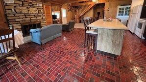 A rustic interior with a stone fireplace, wooden beams, a tiled floor, a sofa, a dining area, and a kitchen with white appliances. - Private, cozy cabin on 13 acres with two ponds (Bloomfield)