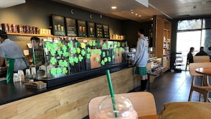 Interior de una cafetería Starbucks con un mostrador de madera, una persona con una bebida y otra trabajando detrás del mostrador.