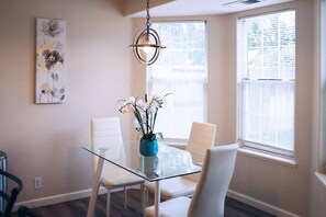Un coin repas avec une table en verre, des chaises blanches, un vase bleu rempli de fleurs et un luminaire suspendu.
