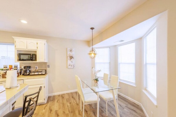 A modern kitchen with white cabinetry, a dining area with a glass table and chairs, and large windows providing natural light.