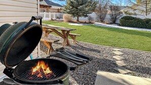 Ein Grill mit Zange und Deckel, ein Picknicktisch und eine Rasenfläche mit Zaun und Bäumen.