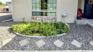 Einbungalow mit Veranda, Steinunterbau und kleinem Garten mit Kies und Pflanzen.