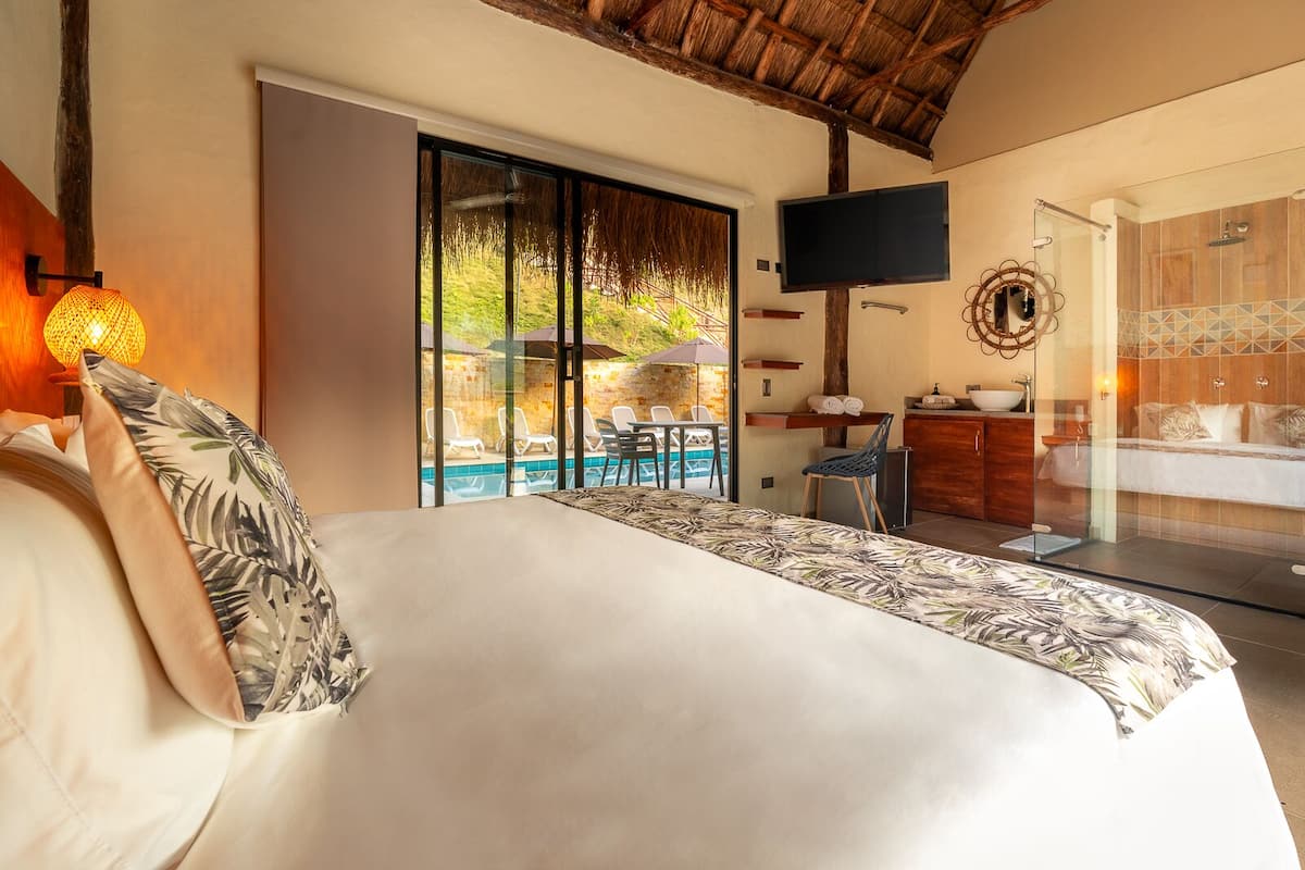 Un dormitorio con una cama grande, un televisor montado en la pared, vista a una piscina y un baño con bañera y lavamanos.