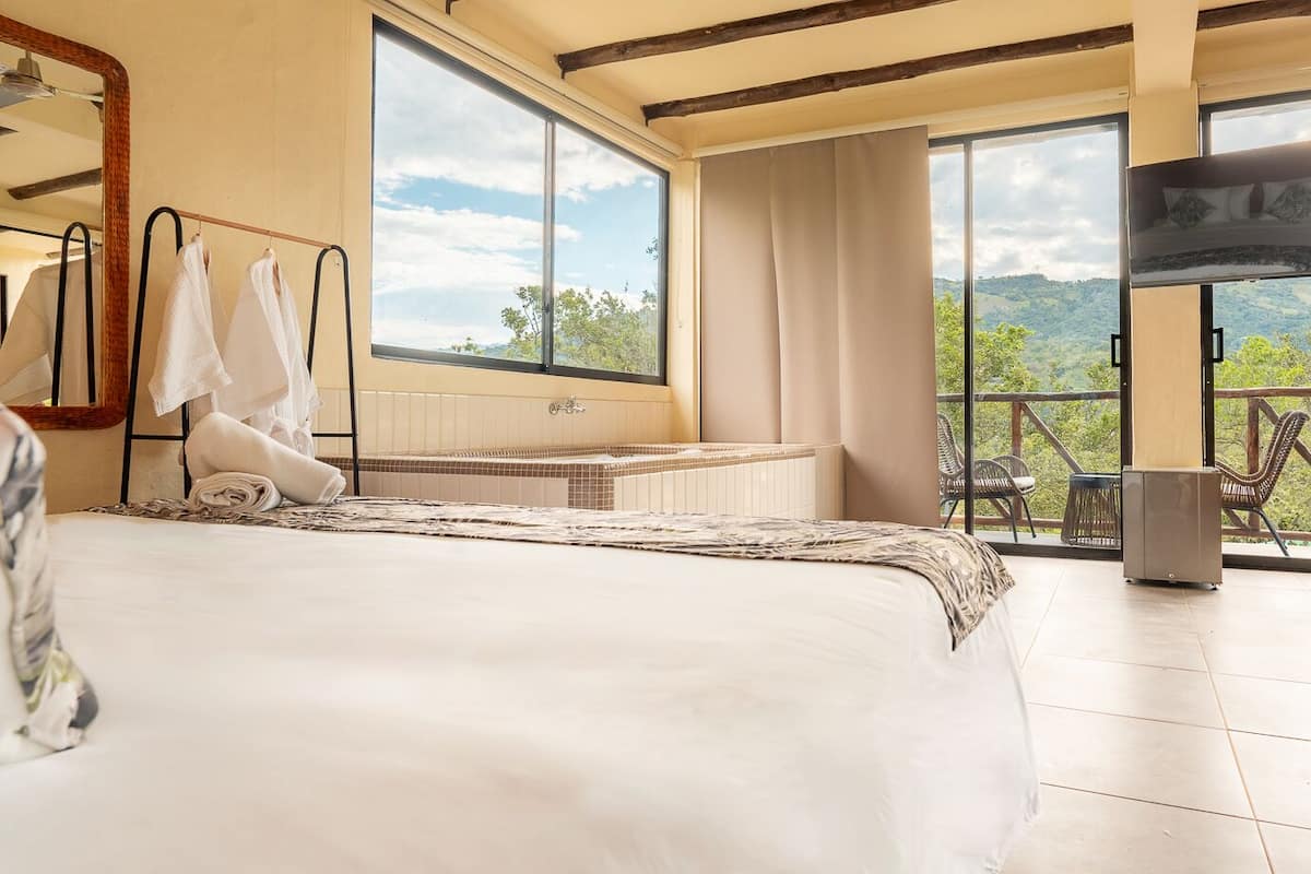 Habitación de hotel con una cama grande, una bañera, un televisor y un balcón con vistas a las montañas.