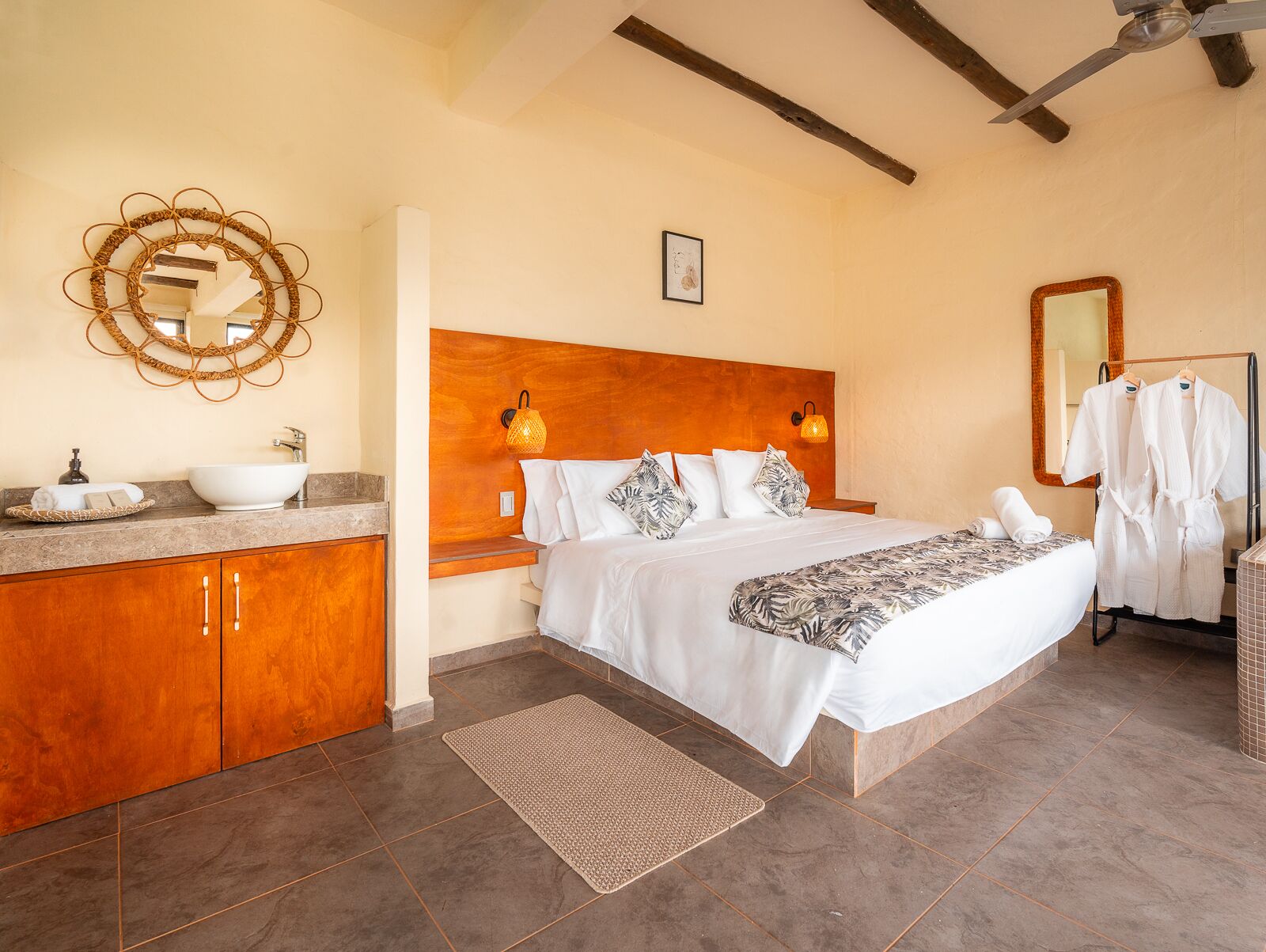 Un dormitorio con cabecera de madera, una cama grande con ropa blanca, una mesita de noche con lámpara, un baño con lavabo y espejo, y un adorno de pared tejido.
