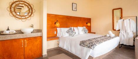 Un dormitorio con cabecera de madera, una cama grande con ropa blanca, una mesita de noche con lámpara, un baño con lavabo y espejo, y un adorno de pared tejido.