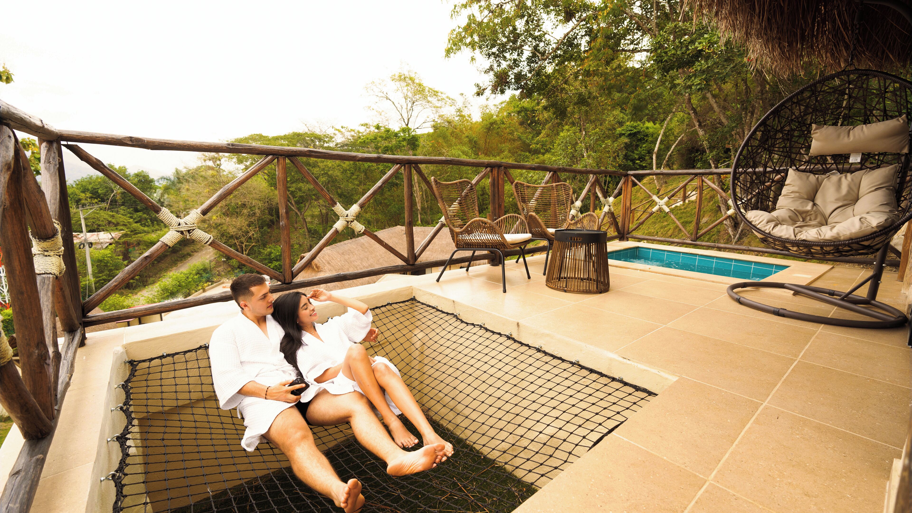 Una pareja descansando en una hamaca junto a una piscina, rodeada de mobiliario de mimbre y una terraza de madera.