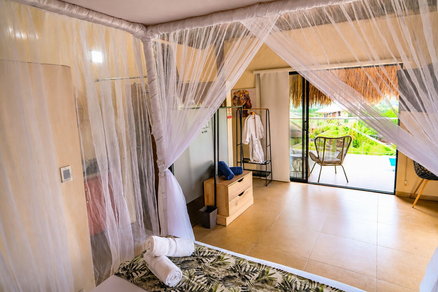 A room with a bed, a wooden chest, a robe rack, and a view of a balcony with chairs.
