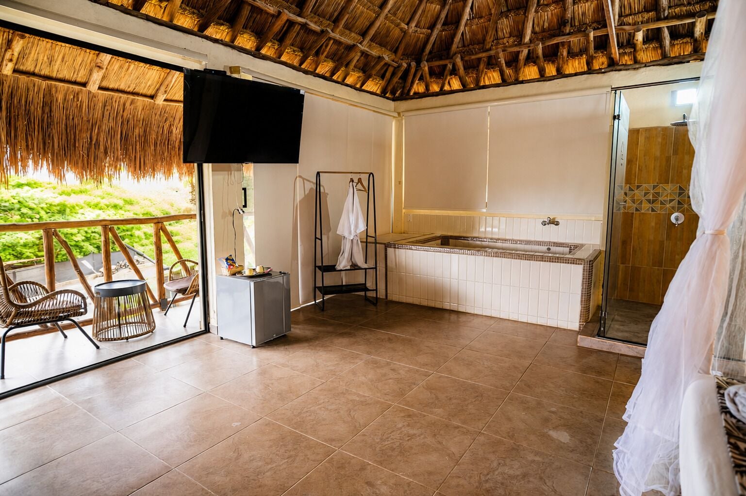 Una habitación con techo de paja, un ventanal grande, una bañera, una ducha y una pequeña mesa con una silla.