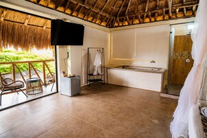 Una habitación con techo de paja, un ventanal grande, una bañera, una ducha y una pequeña mesa con una silla.