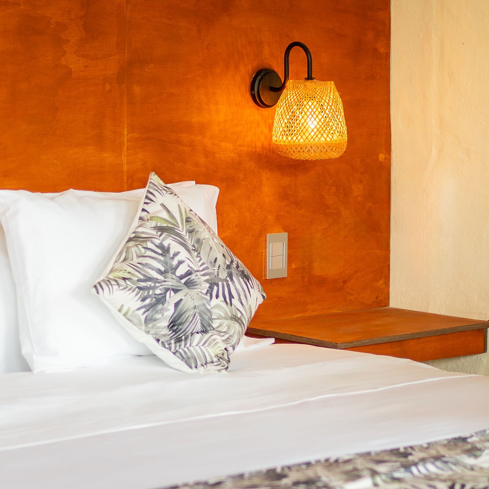 Un lit avec une literie blanche et un coussin orné d’un motif de feuilles tropicales, une lampe fixée au mur et une table de chevet en bois.
