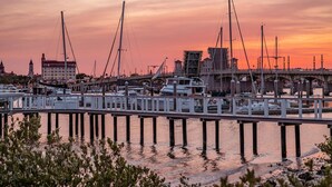 Uma marina com barcos atracados, uma ponte e um edifício com torre do relógio ao fundo.