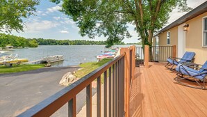 Una terraza de madera con sillas azules, vista a un lago, barcos amarrados y árboles.