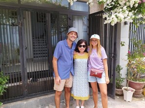 Three people standing in front of a black gate with a white door, surrounded by potted plants and flowers.