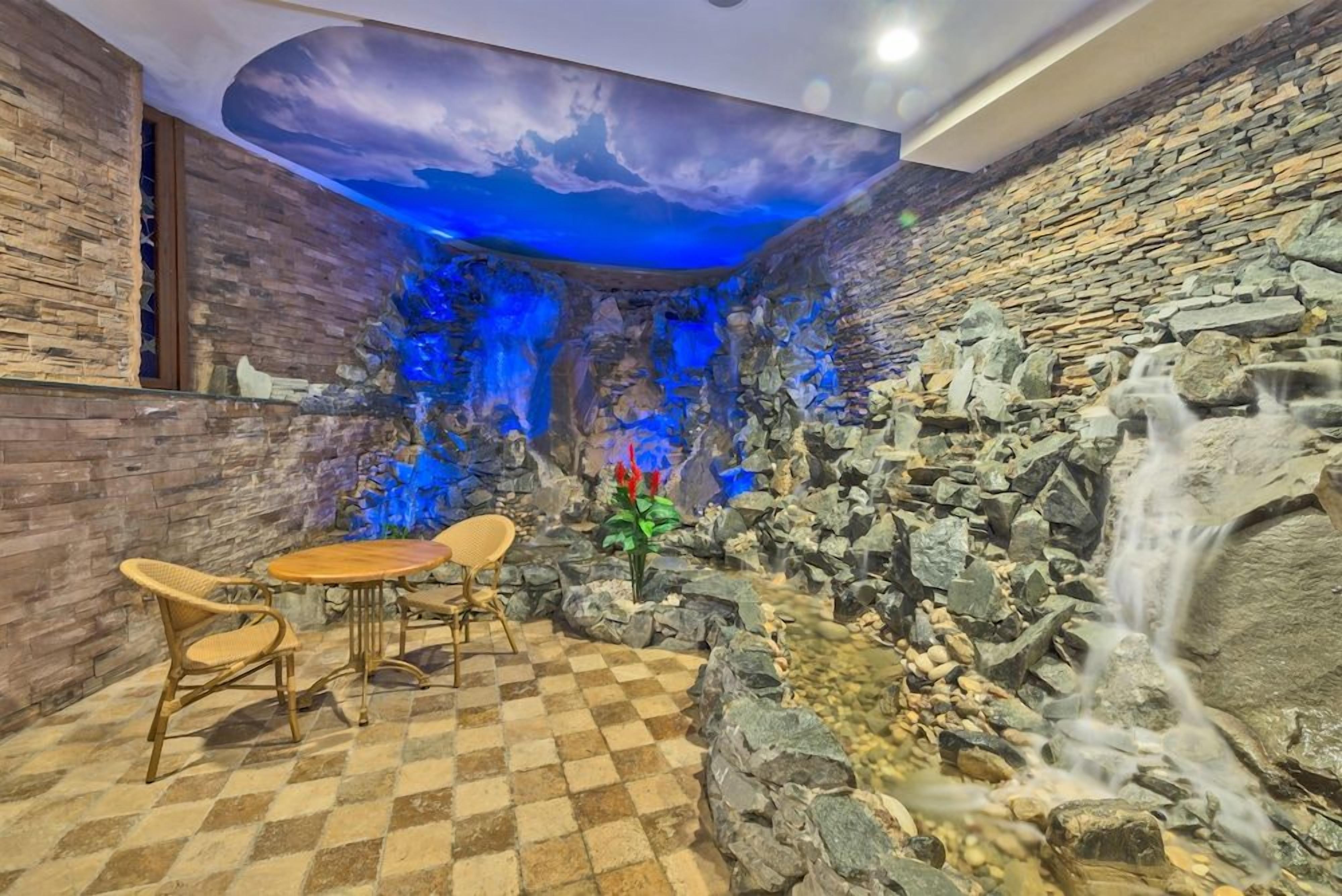 Une pièce dotée d’un sol à carreaux, d’une table entourée de chaises, d’un mur décoratif agrémenté d’une cascade, et d’un plafond orné d’un ciel peint.