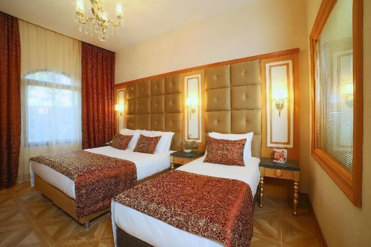 A hotel room with two beds, a large window with curtains, a chandelier, and a wooden headboard.