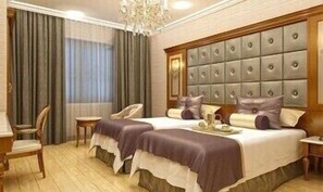 A hotel room with two beds, a large padded headboard, a chandelier, and a wooden floor.