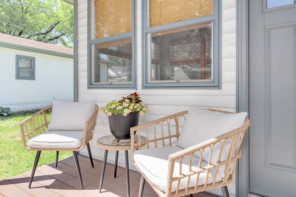 A porch with wicker chairs and a table, a potted plant, and a window with a view of a neighboring house.