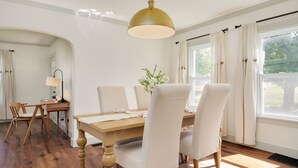 A dining room with a wooden table, white upholstered chairs, a hanging light fixture, and a window with curtains. - Welcome to Casa de Campo! Your Home Away From Home (Saint Joseph)