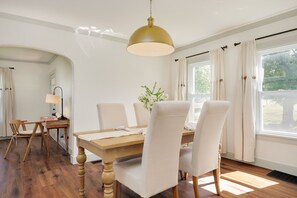 Ein Esszimmer mit Holztisch, weiß gepolsterten Stühlen, einer Hängelampe und einem Fenster mit Vorhängen.