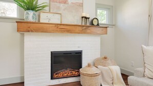 A white brick fireplace with a wooden mantel shelf, a clock, and candles. There are wicker baskets and a throw blanket on the floor. - Welcome to Casa de Campo! Your Home Away From Home (Saint Joseph)