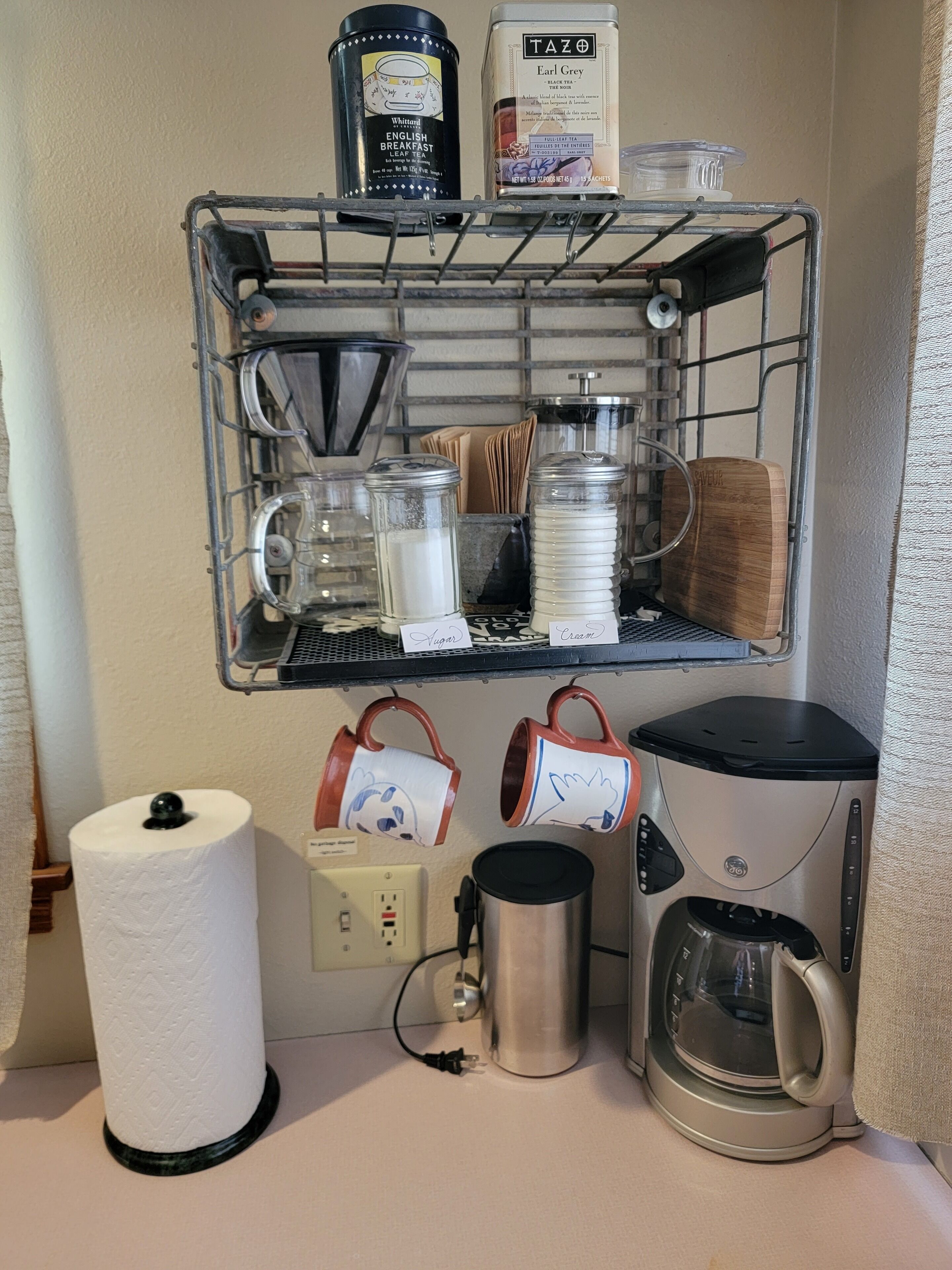 A kitchenette with a coffee maker, a kettle, a tea dispenser, and a shelf with tea bags and a coffee grinder.