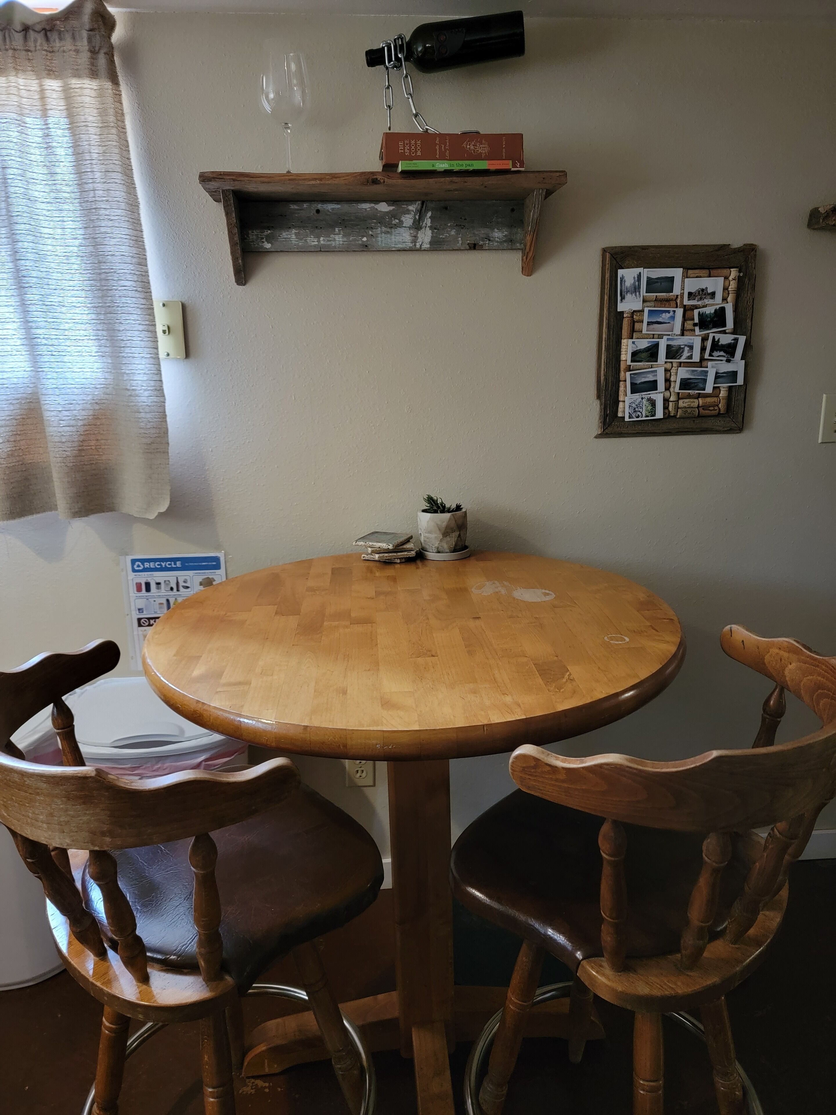 Une table de salle à manger ronde en bois, accompagnée de deux chaises en bois, une étagère avec des livres et une bouteille, et un tableau d’affichage avec des photos accroché au mur.