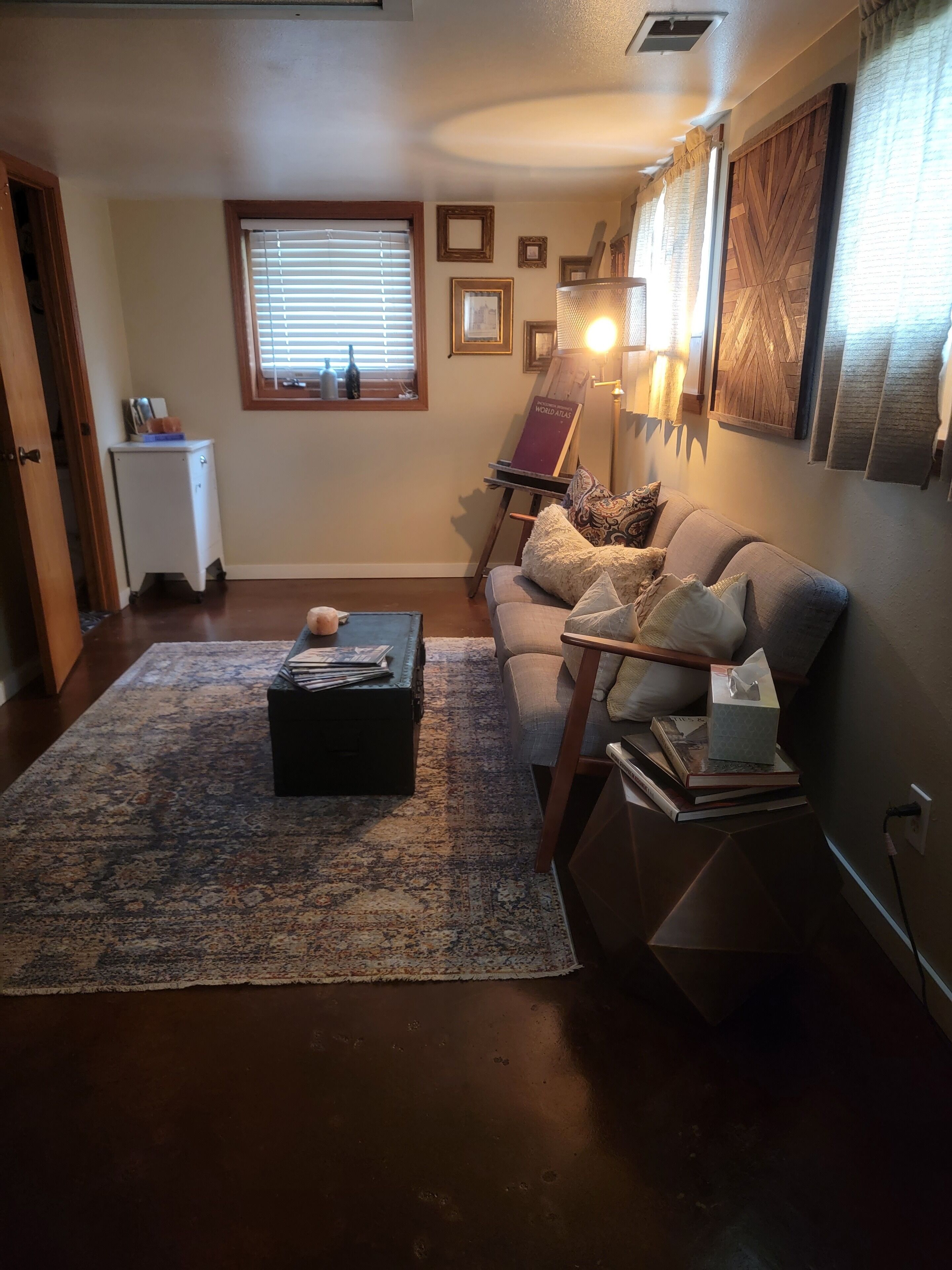 Una sala de estar acogedora con un sofá, una mesa de centro y una alfombra. Se ve una ventana con persianas, una lámpara y cuadros enmarcados en la pared.