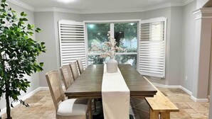 A dining room with a long table, chairs, and a bench. There is a potted plant and a window with blinds. - Premium Heated Pool House with Game Room (Houston)