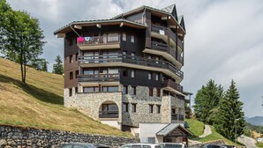 Een meerlaags gebouw met balkons, een gevel van natuursteen en een skipiste op de achtergrond.
