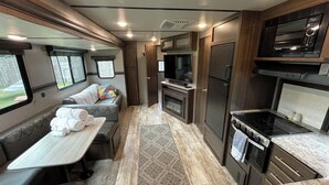 Interior compacto de uma RV com cozinha compacta, área de estar com mesa e almofadas, e uma televisão.