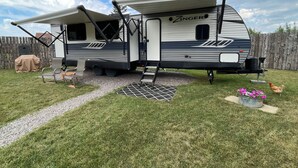 A Zinger RV with a covered patio area, a grill, and a chicken in the yard.
