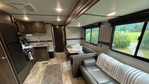 A compact RV interior with a kitchenette, a seating area, and a window offering a view of greenery.