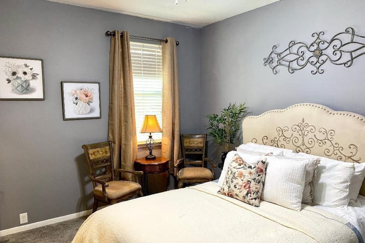 A bedroom with a bed, two chairs, a table with a lamp, and two framed floral artworks on the wall.