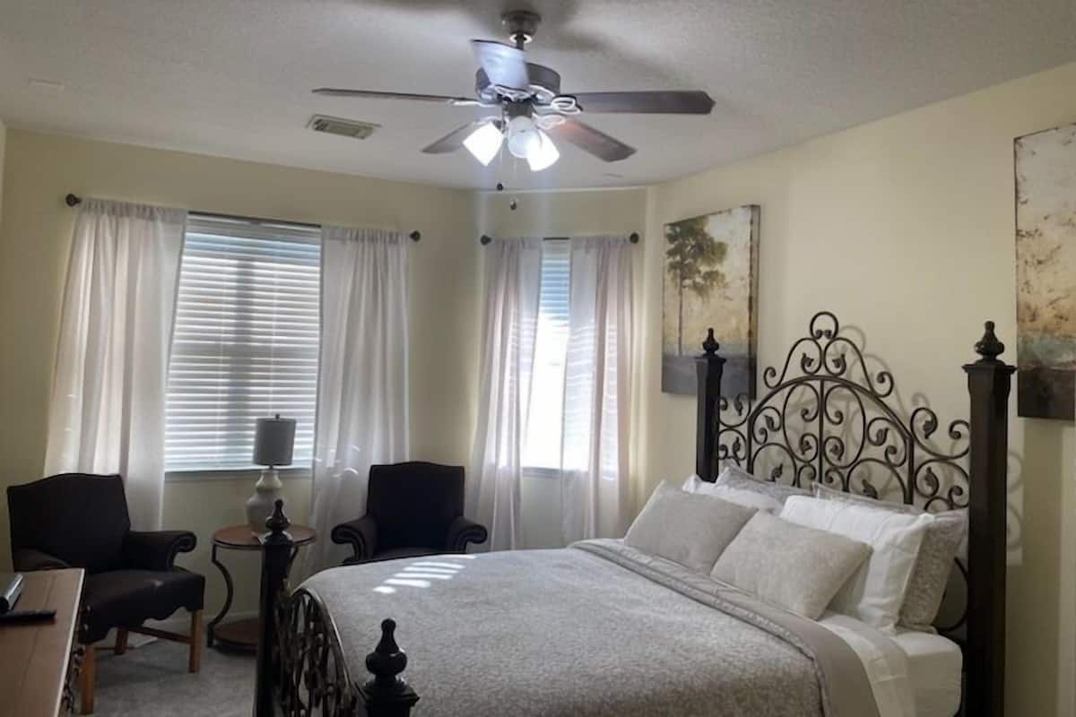 A bedroom with a bed, two chairs, a small table, a ceiling fan, and curtains.