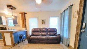 A living room with a brown leather sofa, a wooden coffee table, and a kitchen area with a bar counter and stools. - Cozy private cottage only 2 miles to downtown & ATT center  (San Antonio)