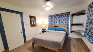 A bedroom with a wooden bed, a nightstand with a lamp, a door with a blue frame, a wall-mounted shelf, and a window with patterned curtains. - Cozy private cottage only 2 miles to downtown & ATT center  (San Antonio)