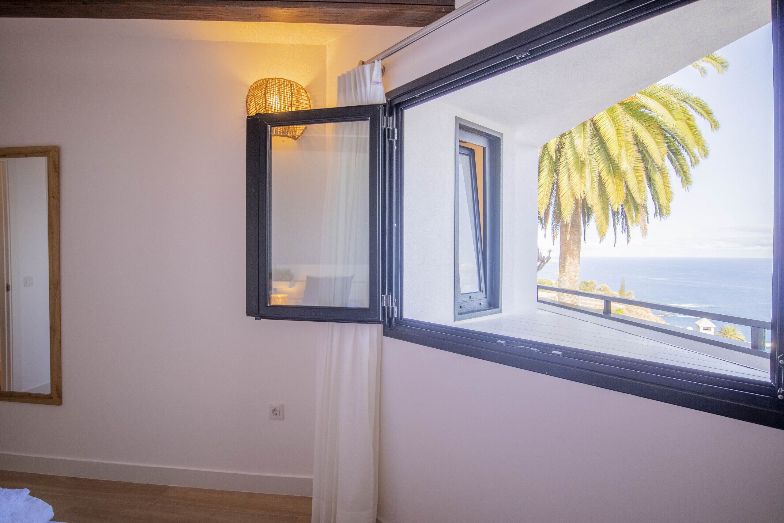 Una habitación con un ventanal grande que da a una vista costera, un espejo y una lámpara de mimbre.