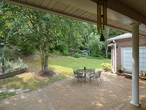 Eine überdachte Terrasse mit Tisch und Stühlen, ein Rasen und ein Backsteinhaus im Hintergrund.
