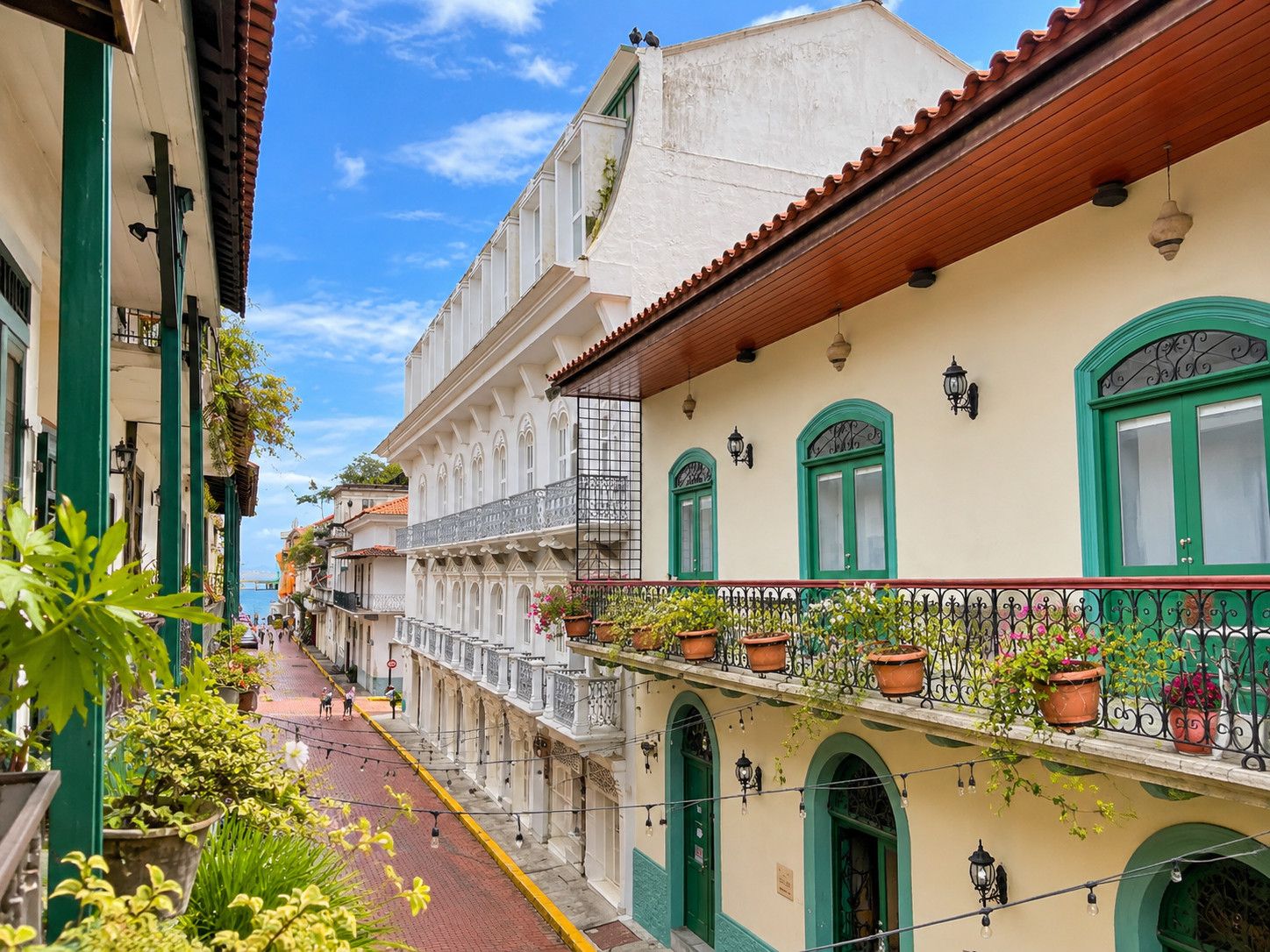 A narrow street lined with buildings featuring balconies with potted plants, a clear sky, and a distant view of the sea.