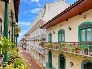 Une rue étroite bordée d’immeubles dotés de balcons ornés de plantes en pot, un ciel dégagé et une vue lointaine sur la mer.