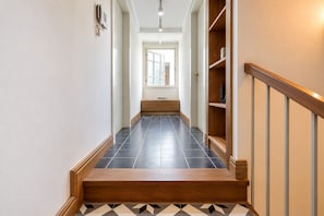 Un pasillo con una escalera de madera, suelo de baldosas y una estantería.