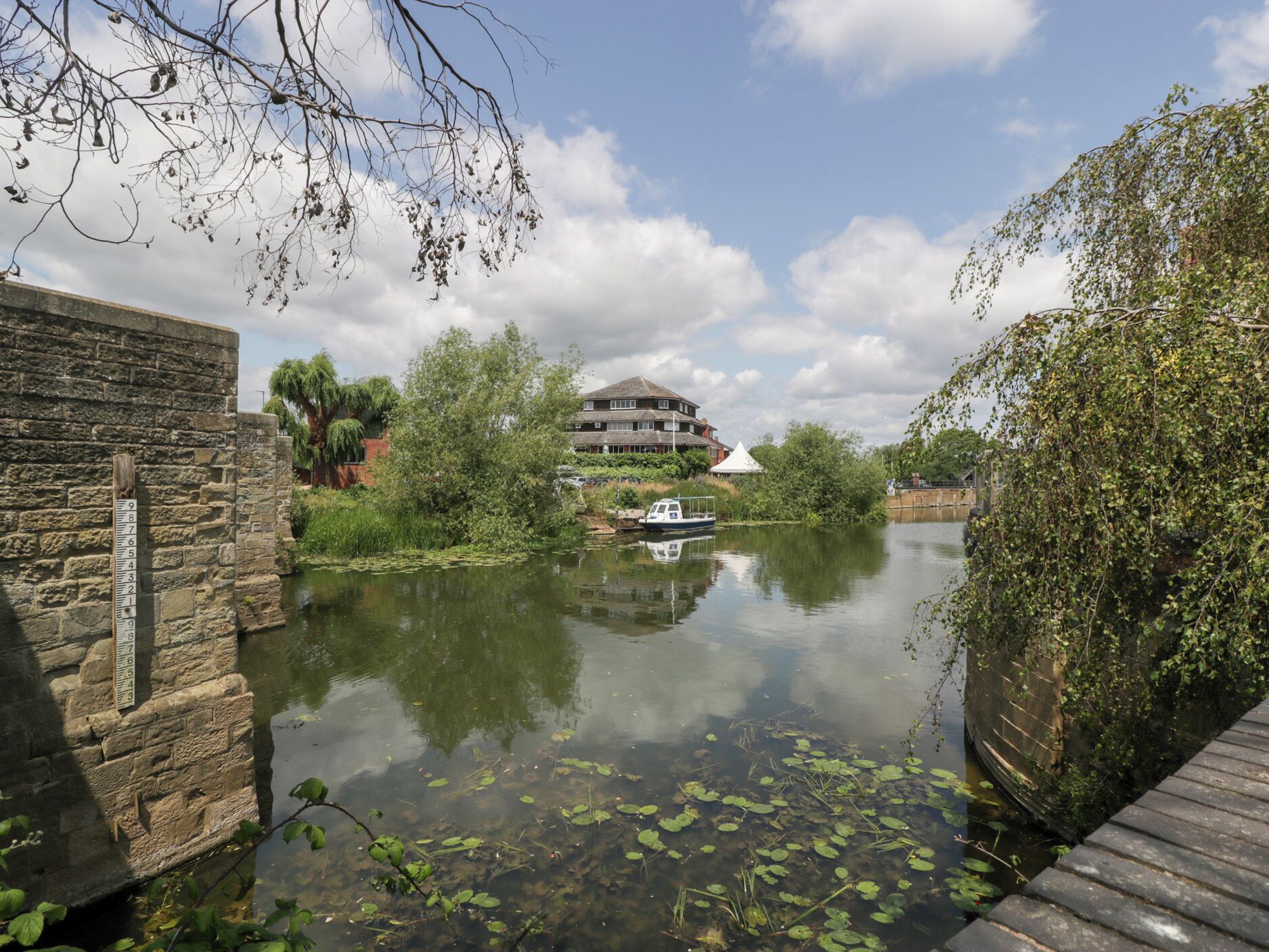A riverside view with a stone wall, a wooden dock, and a building with multiple windows and a triangular roof.
