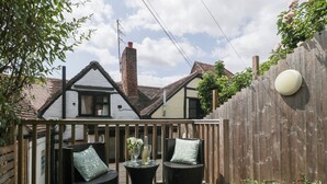 A wooden deck with two wicker chairs and a small table, a wooden fence, and a house with a pitched roof in the background. - Avonmede (Tewkesbury)