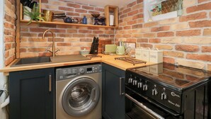 A compact kitchen with a brick wall, a washing machine, a stove, and shelves with kitchenware. - Avonmede (Tewkesbury)