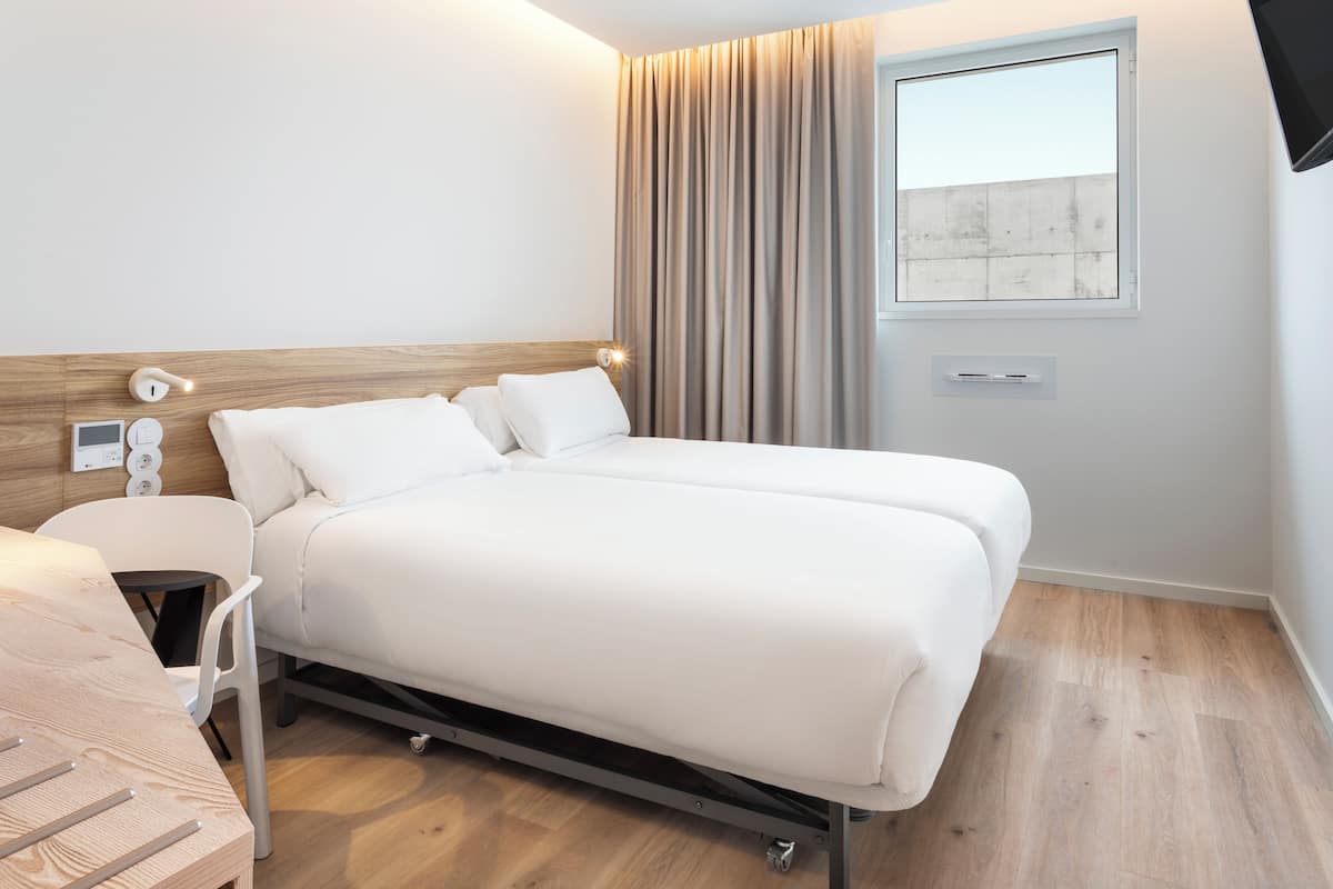Une chambre d’hôtel moderne avec une tête de lit en bois, un lit recouvert de draps blancs, une table de chevet et une fenêtre avec des rideaux.