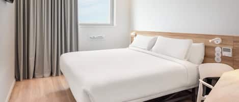 Una habitación de hotel moderna con una cama grande, cabecera de madera, mesita de noche, una silla, ventana con cortinas y una lámpara fijada a la pared.