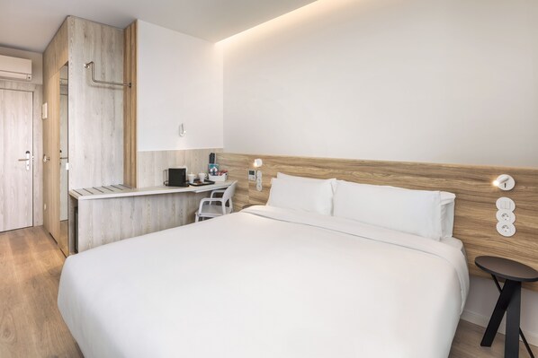 Une chambre d’hôtel moderne avec une tête de lit en bois, un lit recouvert de draps blancs, un petit bureau avec une chaise et une table de chevet avec une lampe.