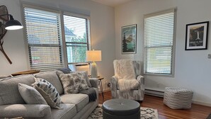 A living room with a grey sofa, a fluffy armchair, a round ottoman, and a patterned rug. There are two windows with blinds, a lamp, and framed pictures on the walls. - Downtown Snoqualmie Home - Hot Tub, Fully Fenced Yard - Pet Friendly! (Snoqualmie)
